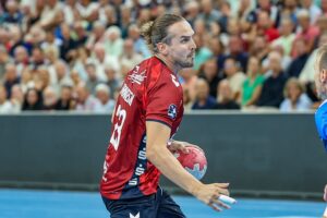 Ein männlicher Handballspieler in einem roten Trikot hält einen Ball und bereitet sich darauf vor, ihn während eines Spiels zu werfen, wobei das Publikum im Hintergrund unscharf zu sehen ist.