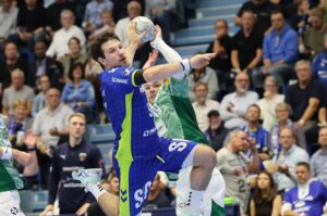 Ein Handballspieler in einem blauen Trikot springt, um den Ball zu schießen, während ein Verteidiger in Grün versucht, ihn zu blockieren. Das Spiel findet in einer Halle statt, in der sich viele Zuschauer aufhalten.