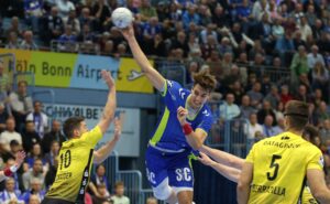 Ein Handballspieler in einem blau-grünen Trikot springt, um den Ball zu schießen, während drei gelb-schwarze Verteidiger versuchen, ihn während eines Spiels zu blockieren, während die Zuschauer im Hintergrund zusehen.