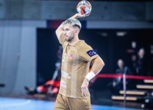 Ein männlicher Handballspieler in einem braunen Trikot hält einen Handball über seinen Kopf und bereitet sich auf den Wurf vor. Der Hintergrund zeigt eine unscharfe Sporthalle mit Menschen und Geräten.