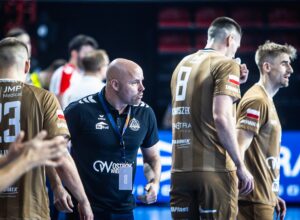 Ein Trainer in einem dunklen Hemd gibt Anweisungen an seine Handballmannschaft, die braune Trikots mit der Nummer 8 trägt. Die Mannschaft scheint sich auf das Spiel in einer Sporthalle zu konzentrieren.