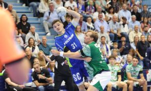 Ein Handballspieler in einem blauen Trikot bereitet sich auf den Wurf vor, während er von einem Gegner in einem grünen Trikot eng verteidigt wird. Die Zuschauer auf den überfüllten Tribünen im Hintergrund schauen aufmerksam zu.