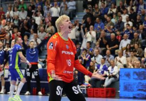 Ein Handballtorwart in einem orangefarbenen Trikot schreit und gestikuliert leidenschaftlich während eines Spiels, während die Zuschauer auf den überfüllten Tribünen im Hintergrund aufmerksam zusehen.