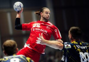 Ein männlicher Handballspieler in einem roten Trikot springt, um den Ball zu werfen, während zwei Gegner in dunklen Trikots versuchen, ihn während eines rasanten Hallenspiels zu blockieren.