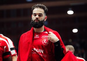 Ein männlicher Handballspieler mit dichtem Bart und kurzen Haaren trägt ein rotes Trikot und ein rotes Handtuch über den Schultern, das er während eines Spiels in der Halle konzentriert hält.