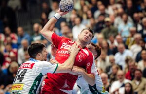 Ein männlicher Handballspieler in einem roten Trikot springt zum Schuss, während zwei Verteidiger in weißen und grünen Trikots versuchen, ihn während eines überfüllten Hallenspiels zu blockieren. Im Hintergrund ist das Publikum zu sehen.