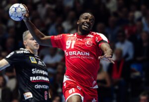 Ein männlicher Handballspieler in einem roten Trikot mit der Nummer 91 springt mit erhobenem Arm, um den Ball zu werfen. Ein anderer Spieler in einem schwarzen Trikot steht hinter ihm. Im Hintergrund ist eine unscharfe Menschenmenge zu sehen.