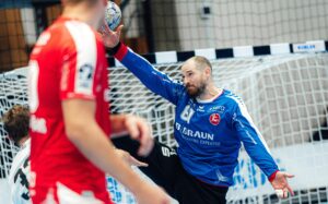 Ein Handballtorwart in einem blauen Trikot springt, um einen Schuss abzublocken, und streckt Arm und Bein in Richtung Ball aus, während im Vordergrund, in der Nähe des Tores, Spieler in roten Trikots zu sehen sind.