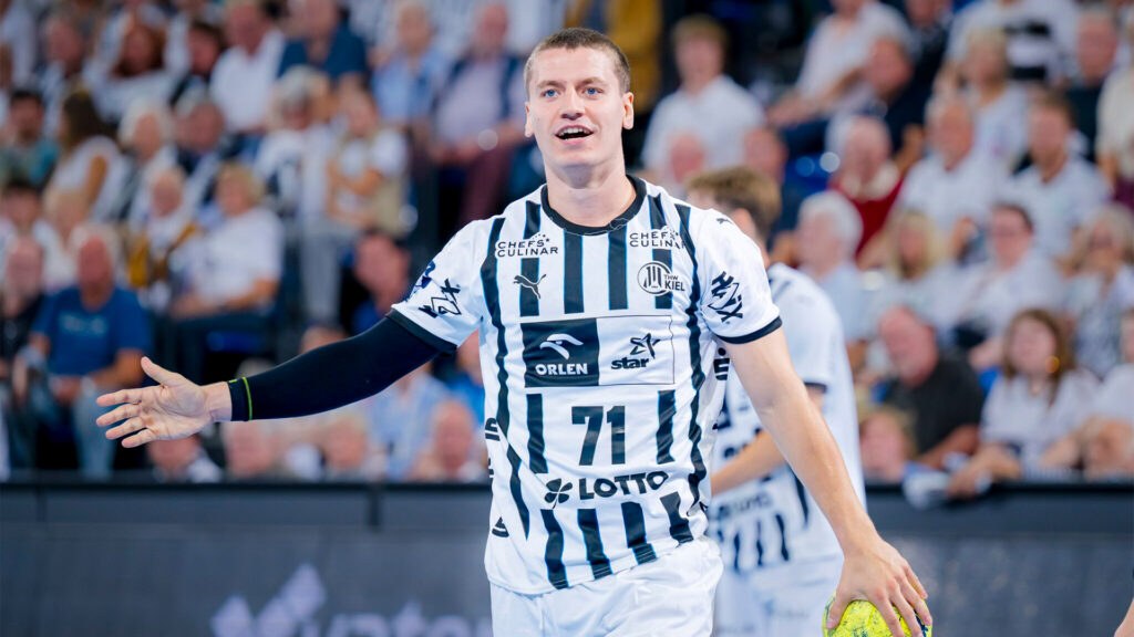 Ein männlicher Handballspieler in einem schwarz-weiß gestreiften Trikot mit der Nummer 71 steht auf dem Spielfeld und lächelt mit einem ausgestreckten Arm. Fans in weißen Hemden füllen den Hintergrund.