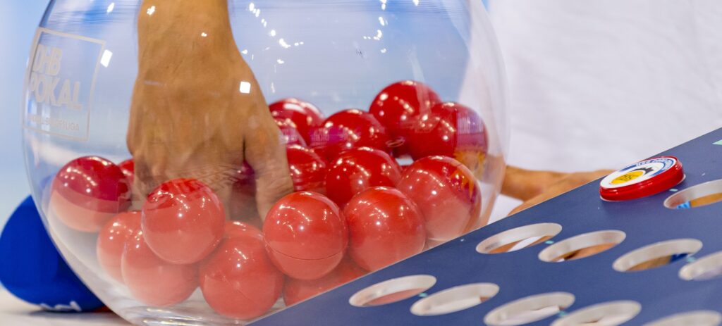 HANDBALL: Pixum Super Cup 2022 Eine Hand greift in eine durchsichtige Schale, die mit roten Lotteriekugeln gefüllt ist. Im Vordergrund befindet sich ein blaues Brett mit kreisförmigen Löchern.
