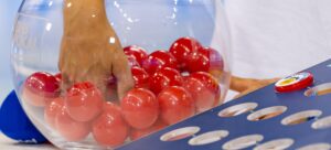 Eine Hand greift in eine durchsichtige Schale, die mit roten Lotteriekugeln gefüllt ist. Im Vordergrund befindet sich ein blaues Brett mit kreisförmigen Löchern.