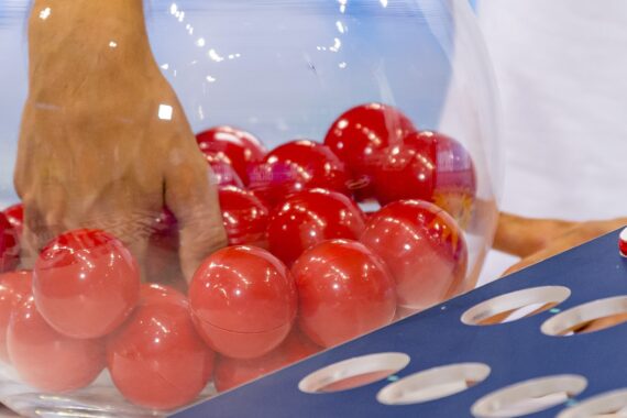 Eine Hand greift in eine durchsichtige Schale, die mit roten Lotteriekugeln gefüllt ist. Im Vordergrund befindet sich ein blaues Brett mit kreisförmigen Löchern.