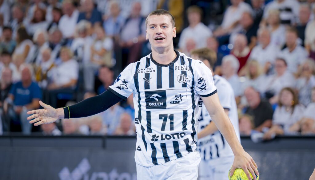 Ein männlicher Handballspieler, der ein weiß-schwarz gestreiftes Trikot mit der Nummer 71 trägt, steht auf dem Spielfeld und hält einen gelben Ball, während im Hintergrund eine unscharfe Zuschauermenge zu sehen ist.