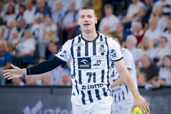 Ein männlicher Handballspieler, der ein weiß-schwarz gestreiftes Trikot mit der Nummer 71 trägt, steht auf dem Spielfeld und hält einen gelben Ball, während im Hintergrund eine unscharfe Zuschauermenge zu sehen ist.