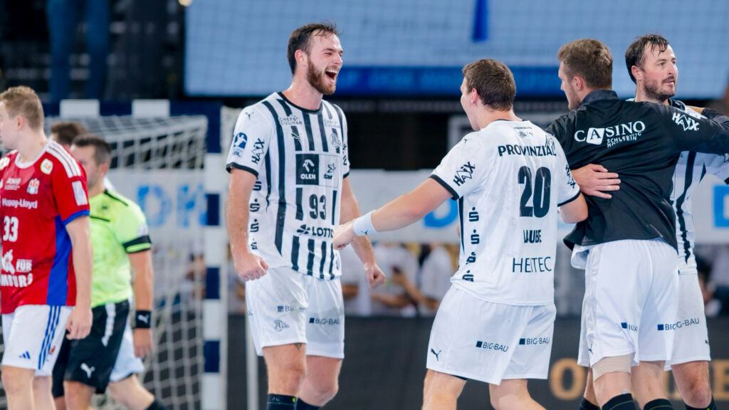 Handballspieler in weißen Trikots feiern auf dem Spielfeld, lächeln und umarmen sich, während eine gegnerische Mannschaft in Rot im Hintergrund davonläuft. Hinter ihnen sind ein Tornetz und eine Anzeigetafel zu sehen.