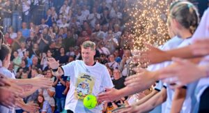 Ein Mann, der einen grünen Fußball in der Hand hält, geht an Menschen vorbei, die sich gegenseitig abklatschen. Er trägt ein T-Shirt mit Grafiken und Text, im Hintergrund ist eine Menschenmenge zu sehen und in der Nähe fliegen Funken.