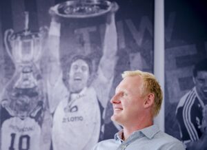 Ein lächelnder Mann mit blondem Haar sitzt im Haus vor einem großen Wandgemälde in Graustufen, das jubelnde Sportler mit einer Trophäe zeigt.