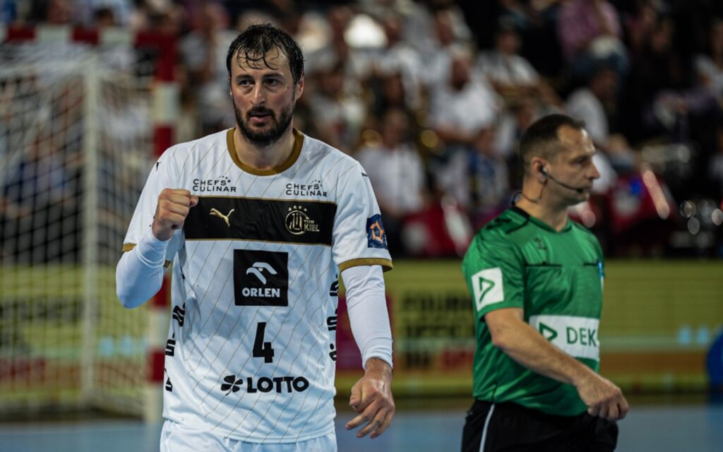 mhb_dule Ein männlicher Handballspieler in einem weißen Trikot mit der Nummer 4 jubelt mit geballter Faust; ein Schiedsrichter in einem grünen Trikot steht neben ihm auf einem Hallenplatz, die Zuschauer sind unscharf im Hintergrund zu sehen.