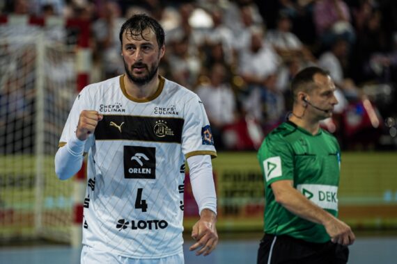 Ein männlicher Handballspieler in einem weißen Trikot mit der Nummer 4 jubelt mit geballter Faust; ein Schiedsrichter in einem grünen Trikot steht neben ihm auf einem Hallenplatz, die Zuschauer sind unscharf im Hintergrund zu sehen.