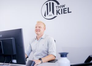 Ein Mann mit kurzen blonden Haaren und einem hellgrauen Hemd sitzt lächelnd an einem Schreibtisch mit Monitor. An der Wand hinter ihm ist das Logo des THW Kiel und die Jahreszahl 1904 zu sehen. Im Vordergrund ist eine Wasserflasche zu sehen.