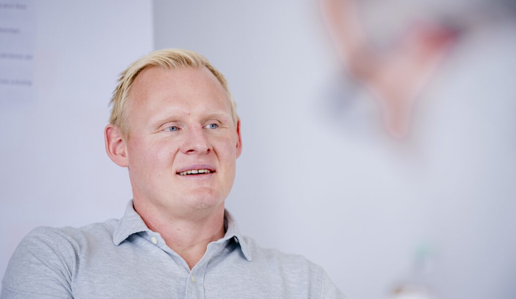 Ein Mann mit kurzen blonden Haaren und einem hellgrauen Hemd mit Kragen lächelt leicht und blickt zur Seite. Der Hintergrund ist unscharf mit weichen Tönen und undeutlichen Formen.