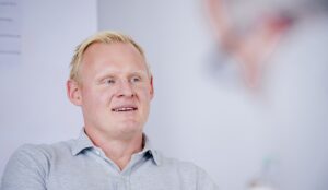 Ein Mann mit kurzen blonden Haaren und einem hellgrauen Hemd mit Kragen lächelt leicht und blickt zur Seite. Der Hintergrund ist unscharf mit weichen Tönen und undeutlichen Formen.