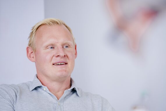 Ein Mann mit kurzen blonden Haaren und einem hellgrauen Hemd mit Kragen lächelt leicht und blickt zur Seite. Der Hintergrund ist unscharf mit weichen Tönen und undeutlichen Formen.