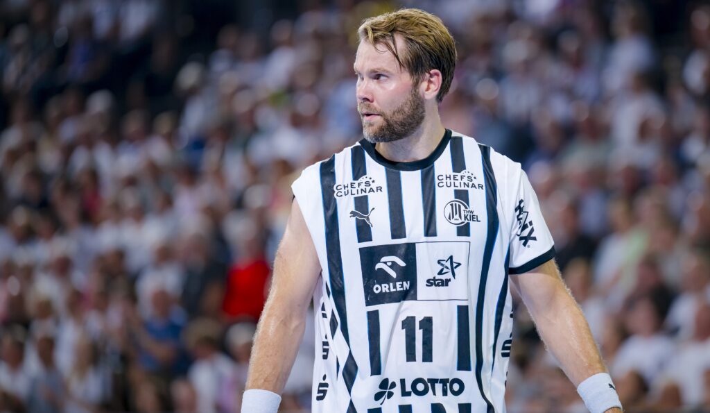 HANDBALL 2025/26: THW Kiel - HSG Wetzlar Ein männlicher Handballspieler, der ein weiß-schwarz gestreiftes THW Kiel-Trikot mit der Nummer 11 trägt, steht auf dem Spielfeld und blickt zur Seite. Im Hintergrund ist eine unscharfe Zuschauermenge während der Handball-Bundesliga Saison 2025/2026 zu sehen.