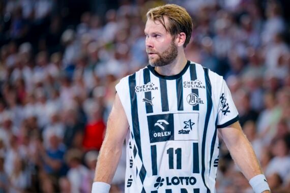 Ein männlicher Handballspieler, der ein weiß-schwarz gestreiftes THW Kiel-Trikot mit der Nummer 11 trägt, steht auf dem Spielfeld und blickt zur Seite. Im Hintergrund ist eine unscharfe Zuschauermenge während der Handball-Bundesliga Saison 2025/2026 zu sehen.