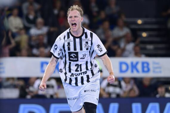 Ein männlicher Handballspieler in einem schwarz-weiß gestreiften Trikot mit der Nummer 21 jubelt leidenschaftlich auf dem Spielfeld, mit offenem Mund und geballten Fäusten. Im unscharfen Hintergrund sind Zuschauer zu sehen.