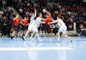 Ein Handballspieler in einem roten Trikot springt zum Schuss, während er von Gegnern in weißen Trikots auf einem Hallenplatz umgeben ist. Die Aktion findet vor einem vollen Publikum auf der Tribüne statt.