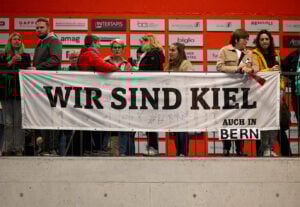 Eine Gruppe von Personen steht hinter einem Geländer und hält ein grosses weisses Transparent mit der Aufschrift "WIR SIND KIEL AUCH IN BERN" vor einer roten Wand mit Sponsorenlogos.