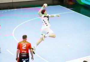 Ein Handballspieler in einem weißen Trikot springt in die Luft, um den Ball in Richtung Tor zu schießen, während ein Verteidiger in einem orange-schwarzen Trikot in der Nähe auf dem blauen Spielfeld steht.