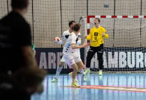 Ein Handballtorwart in einem gelben Trikot jubelt nach einer Parade, während zwei Spieler in Weiß während eines Spiels in einer Halle in der Nähe des Tores reagieren. Im Vordergrund ist eine Teilansicht eines anderen Spielers zu sehen.