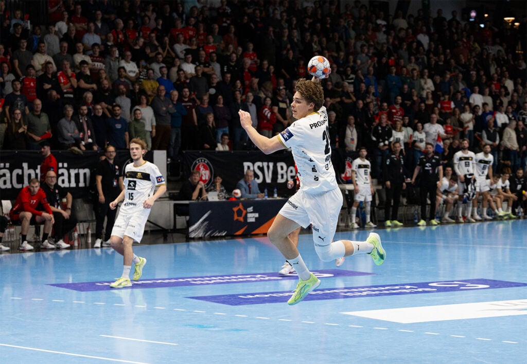 251111_Bern_THW_Rohwer Ein männlicher Handballspieler in einem weißen Trikot springt in der Luft, um den Ball in Richtung Tor zu werfen, während seine Mannschaftskameraden und ein großes Publikum in einer hell erleuchteten Halle zusehen.