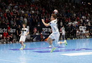 Ein männlicher Handballspieler in einem weißen Trikot springt in der Luft, um den Ball in Richtung Tor zu werfen, während seine Mannschaftskameraden und ein großes Publikum in einer hell erleuchteten Halle zusehen.