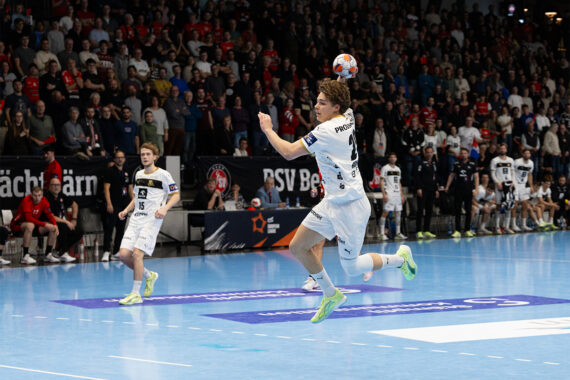 Ein männlicher Handballspieler in einem weißen Trikot springt in der Luft, um den Ball in Richtung Tor zu werfen, während seine Mannschaftskameraden und ein großes Publikum in einer hell erleuchteten Halle zusehen.