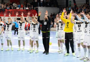 Eine Herren-Handballmannschaft in weißen Trikots steht auf einem Hallenplatz und hebt unisono die Arme, um die Zuschauer zu begrüßen oder ihnen zu danken. Die Zuschauer schauen von den Tribünen im Hintergrund zu.