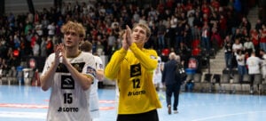 Zwei männliche Handballspieler in Uniform, einer in Weiß (15) und einer in Gelb (12), applaudieren beim Verlassen eines Hallenplatzes. Im Hintergrund ist eine unscharfe Menge von Zuschauern zu sehen.