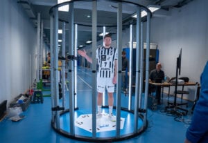 Ein männlicher Fußballspieler in einem schwarz-weiß gestreiften Trikot steht in einem 3D-Scan-Rig mit vertikalen Lichtern, während im Hintergrund einer Sportanlage zwei Personen an Computern sitzen.