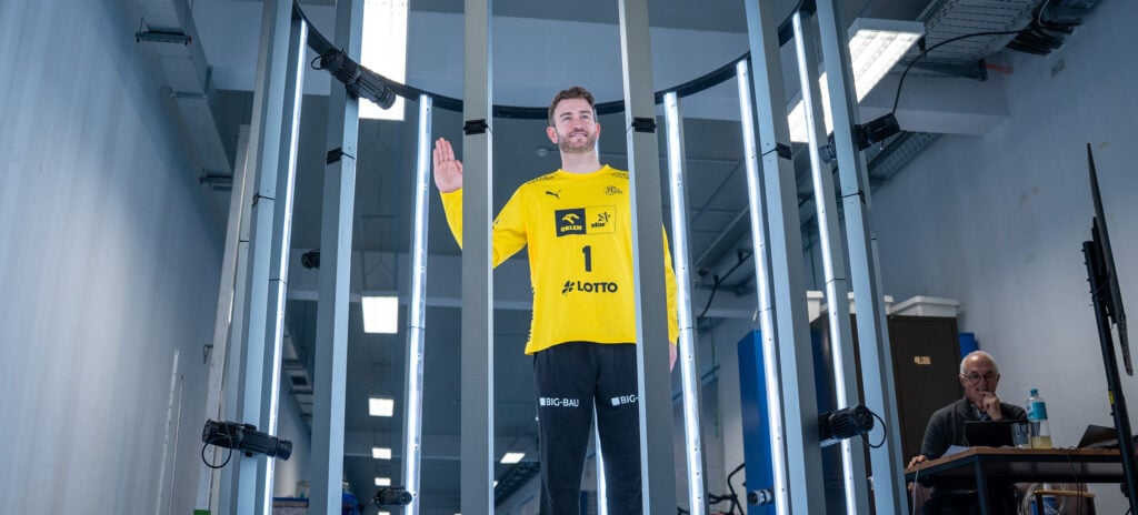 Ein Mann in einem gelben Sporttrikot steht in einem zylindrischen Scannergerät und hält eine Hand hoch. Der Raum ist hell erleuchtet, die Kabel liegen frei und im Hintergrund sitzt eine Person an einem Schreibtisch.