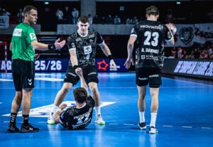 Drei männliche Handballspieler und ein Schiedsrichter befinden sich auf einem Hallenplatz. Ein Spieler sitzt auf dem Boden, während ein Mannschaftskamerad ihm aufhilft, und der Schiedsrichter gestikuliert in der Nähe. Alle Spieler tragen schwarze Trikots, der Schiedsrichter ist grün gekleidet.