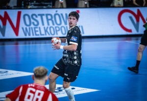 Ein männlicher Handballspieler in einem schwarzen Trikot hält den Ball und bereitet sich auf den Wurf vor, während ein anderer Spieler in einem rot-weißen Trikot ihm gegenübersteht. Der Hintergrund zeigt ein blaues Spielfeld und ein Schild mit der Aufschrift Ostrów Wielkopolski.