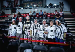 Eine Gruppe von sieben Fans in schwarz-weiß gestreiften Trikots und zebragemusterten Hosen posiert und lächelt in einer Sporthalle, hinter der leere rote und schwarze Sitze stehen.