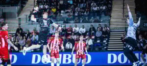 Ein Handballspieler in einem schwarzen Trikot springt in die Luft, um zu schießen, während ein Torwart in Grau sich darauf vorbereitet, den Ball abzufangen. Mehrere Spieler in Rot und Fans im Stadion beobachten das Geschehen.