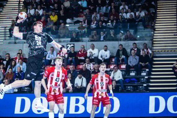 Ein Handballspieler in einem schwarzen Trikot springt in die Luft, um zu schießen, während ein Torwart in Grau sich darauf vorbereitet, den Ball abzufangen. Mehrere Spieler in Rot und Fans im Stadion beobachten das Geschehen.