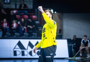 Ein Handballtorwart in einem leuchtend gelben Trikot hebt jubelnd die Faust auf dem Spielfeld, während im Hintergrund Zuschauer sitzen.