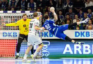 Ein Handballspieler in einem blauen Trikot springt in der Luft zur Seite, um einen Schuss abzugeben, während sich zwei Verteidiger in Weiß und ein gelb gekleideter Torwart darauf vorbereiten, ihn abzuwehren. Eine Menschenmenge schaut von der Tribüne im Hintergrund zu.