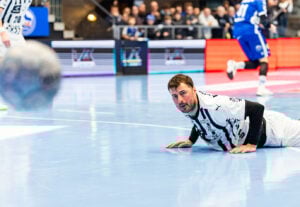 Ein männlicher Handballspieler in einem weiß-schwarzen Trikot liegt auf dem Spielfeld und schaut auf den Ball, der im Vordergrund verschwommen ist. Im Hintergrund sind andere Spieler und Zuschauer zu sehen.