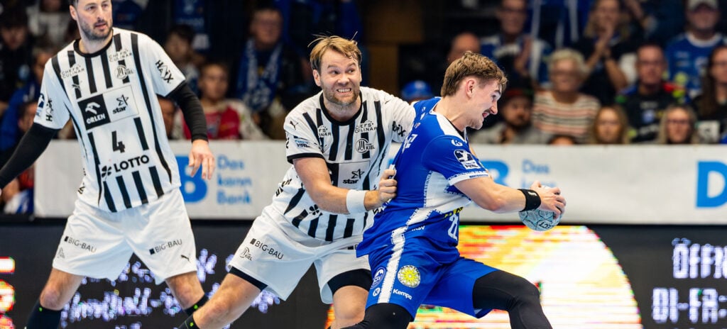 251128_EIS_THW_Overby_Titel Zwei Handballspieler in schwarz-weißen Trikots verteidigen sich gegen einen Spieler in einem blauen Trikot, der den Ball hält, während im Hintergrund eine Zuschauermenge zusieht.